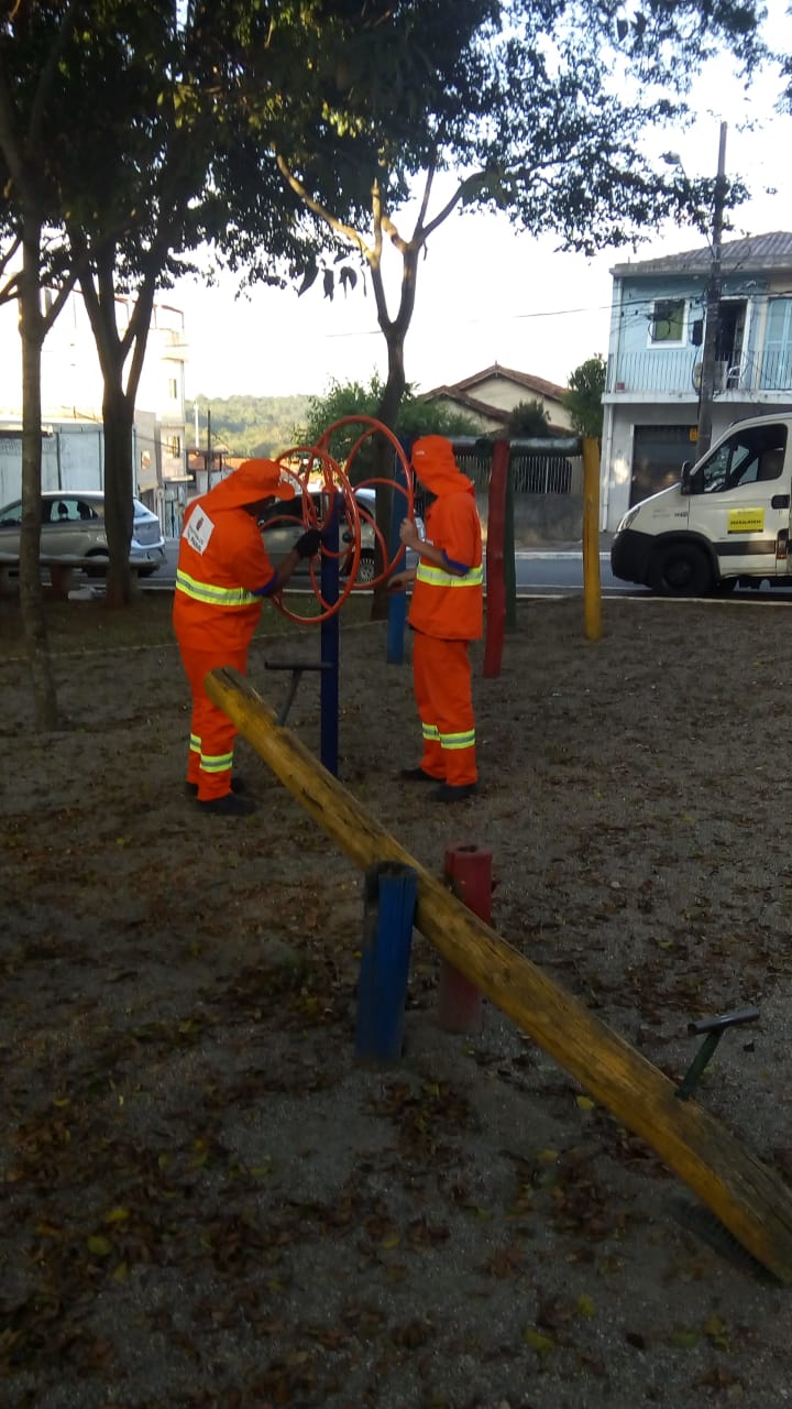 Fotografia de uma praça onde se encontra dois funcionários da prefeitura fazendo a manutenção de aparelhos de exercícios 